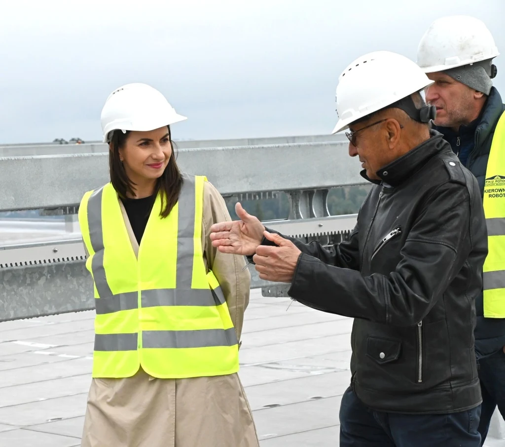 Trzy osoby w kaskach ochronnych i kamizelkach odblaskowych prowadzą rozmowę na placu budowy, w tle widoczne są metalowe konstrukcje i niebo.