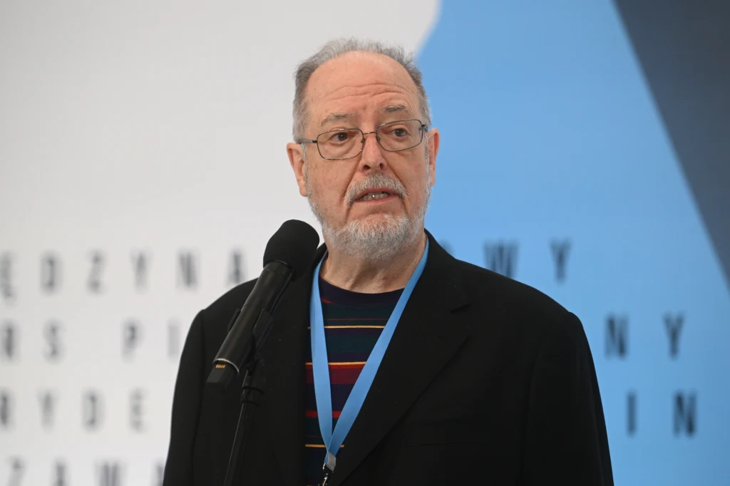 Garrick Ohlsson, przewodniczący jury Mężczyzna w okularach i z siwiejącą brodą mówi do mikrofonu, ubrany w czarną marynarkę i kolorowy sweter, na szyi zawieszony ma niebieski identyfikator; w tle rozmyty napis na ścianie.