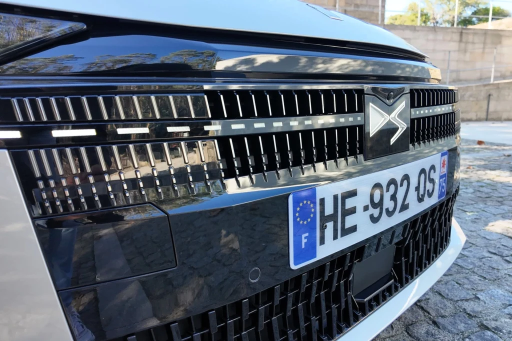 Zbliżenie na przedni grill samochodu marki DS z widoczną tablicą rejestracyjną oraz charakterystycznym logotypem, ostrymi liniami i detalami wykończeniowymi.