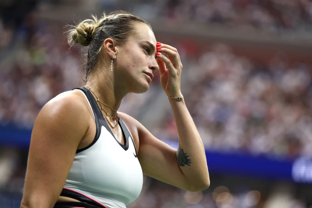 Aryna Sabalenka Zawodniczka tenisowa z wyrazem skupienia na twarzy, ubrana w biały top sportowy z czarnymi dodatkami, jedna ręka przy czole, w tle rozmyta publiczność na trybunach kortu tenisowego.
