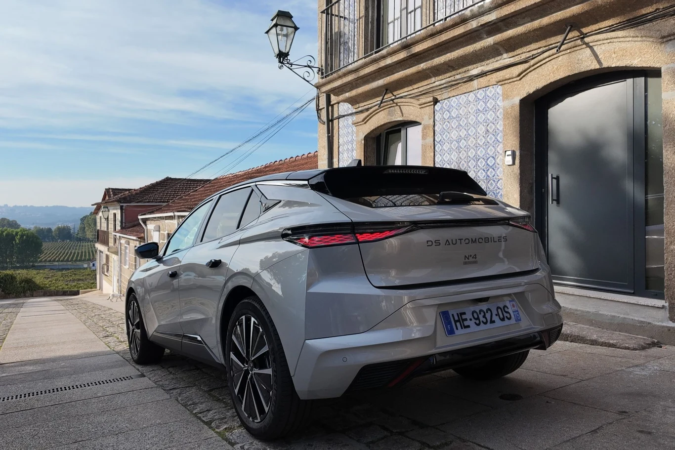 DS N4 z hybrydą plug-in ma przejechać na prądzie do 81 km. Srebrny samochód osobowy DS Automobiles zaparkowany na brukowanej ulicy przy kamienicy z balkonem, w tle widoczne są zielone pola i zabudowania.
