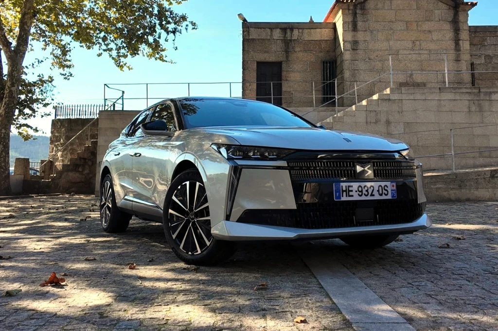 Nowoczesny samochód osobowy w srebrnym kolorze stoi zaparkowany na kamiennym placu przy zabytkowym budynku z kamienia. Po bokach widoczne są nieliczne drzewa, rzucające lekkie cienie na auto.