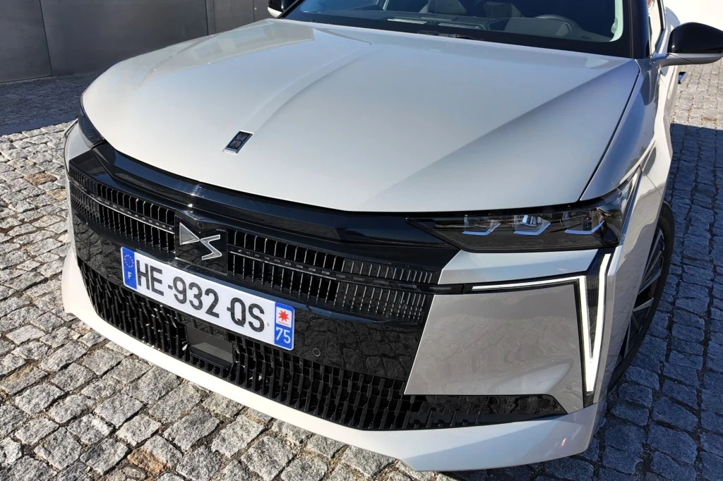 Przód białego samochodu DS z charakterystycznym czarnym grillem, nowoczesnym wzornictwem świateł LED i francuską tablicą rejestracyjną, samochód stoi na kamiennej nawierzchni.