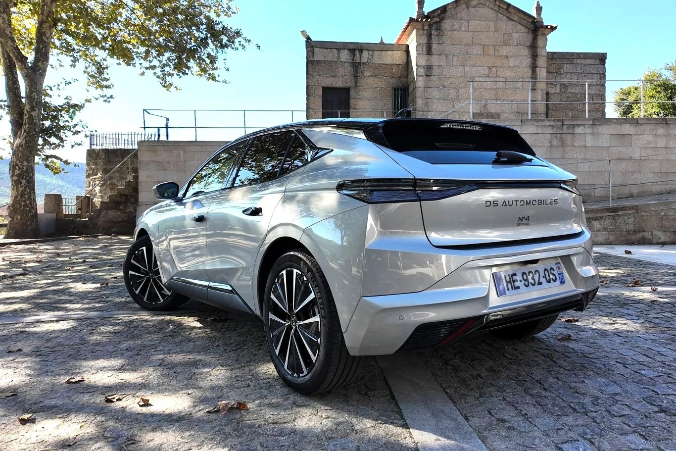 DS N4 dostępne będzie w trzech wariantach napędowych: Hybrid o mocy 145 KM, Plug-in Hybrid o mocy 225 i E-Tense generującej 213 KM. Srebrny samochód marki DS Automobiles zaparkowany na brukowanym placu, w tle kamienny budynek oraz drzewa dające cień.