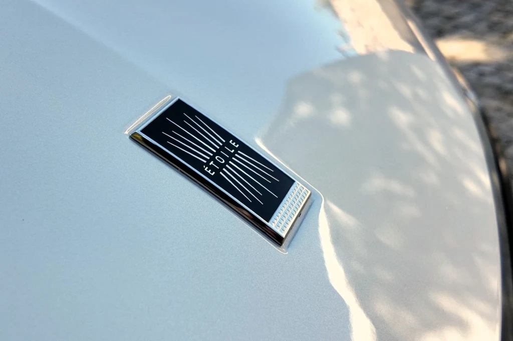 Zbliżenie na srebrny emblemat z napisem ÉTOILE, umieszczony na jasnej masce samochodu, z delikatnymi odbiciami światła wokół znaczka.