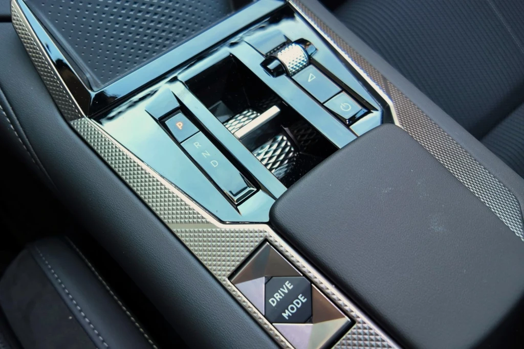 Panel sterowania skrzyni biegów w nowoczesnym samochodzie z widocznymi przyciskami i pokrętłem trybu jazdy, wykończony metalicznymi i czarnymi elementami.