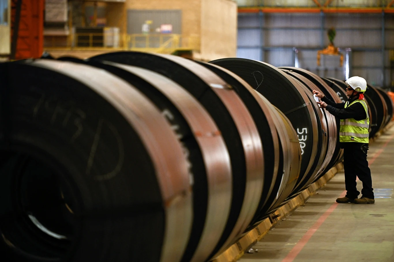 Niemiecka stal coraz śmielej wchodzi na polski rynek pracownik w kamizelce odblaskowej i kasku ochronnym kontroluje duże zwoje blachy stalowej ustawione w rzędzie w hali przemysłowej