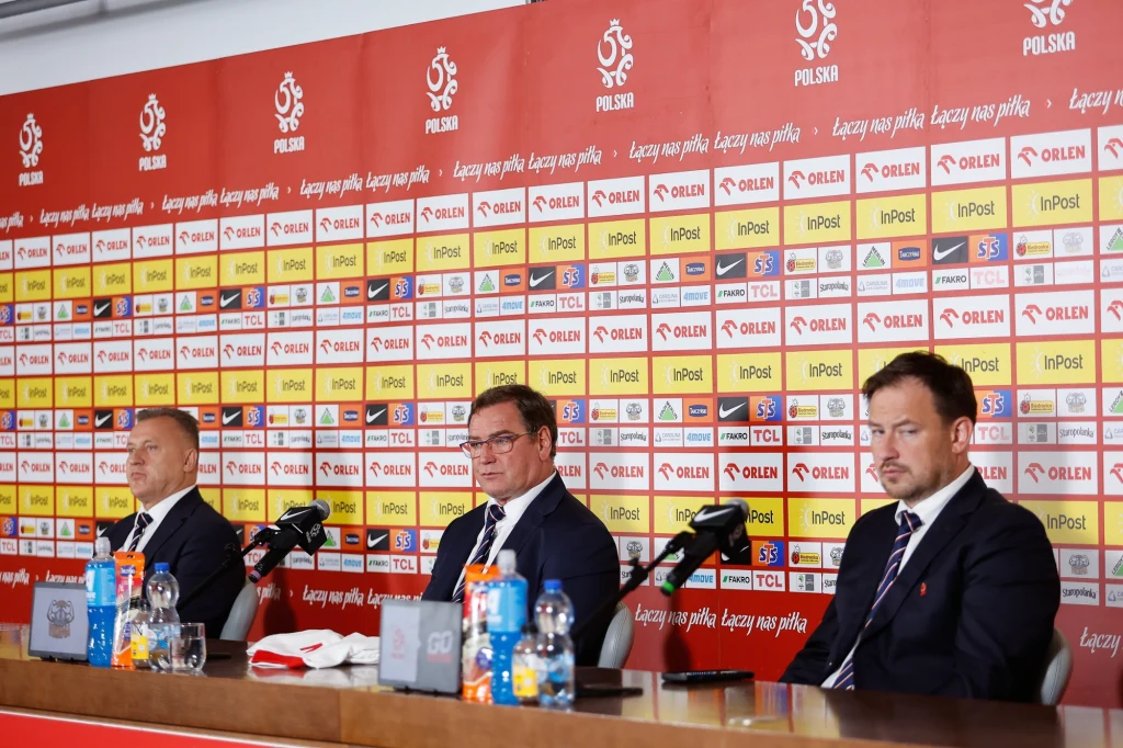 Cezary Kulesza, Jan Urban i Łukasz Wachowski podczas prezentacji nowego selekcjonera reprezentacji Polski Trzech mężczyzn w garniturach siedzi przy stole podczas konferencji prasowej, na stole stoją mikrofony, napoje i dokumenty, za nimi znajduje się ścianka z logotypami sponsorów oraz polskiej federacji piłkarskiej.