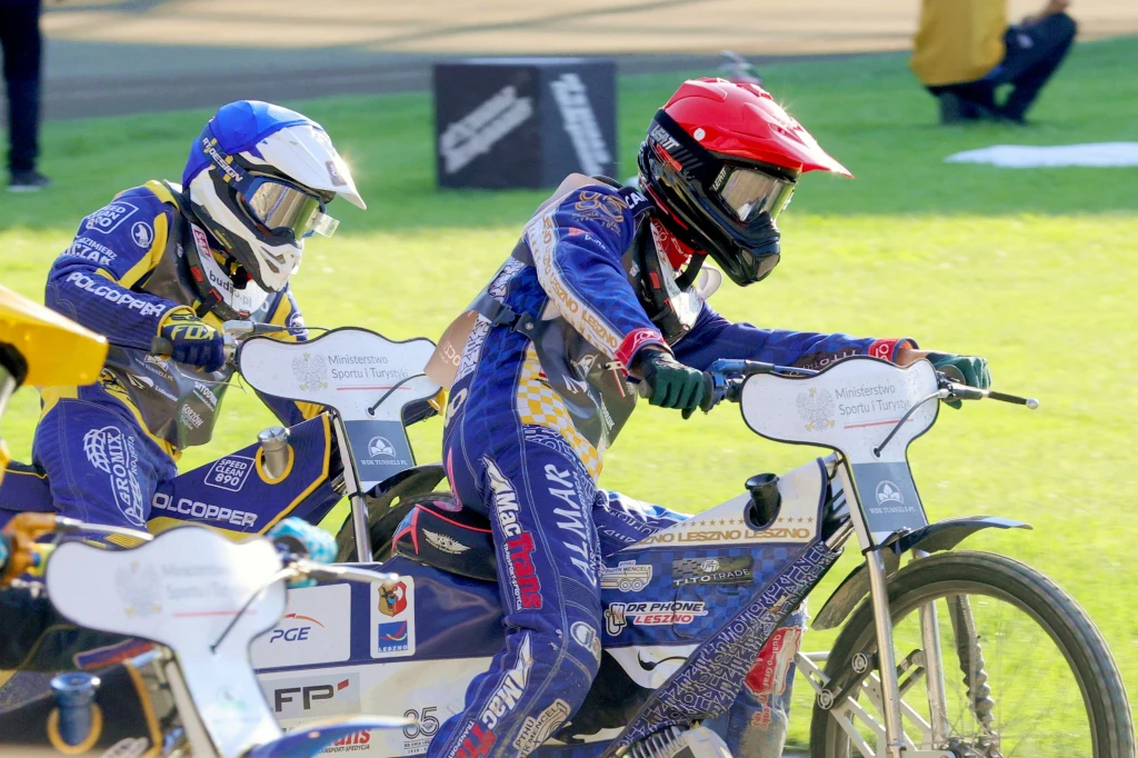 Antoni Mencel przed Nazarem Parnickim. Zawodnicy żużlowi w trakcie wyścigu, ubrani w pełne kombinezony ochronne i kaski, jadący na motocyklach bez hamulców na torze żużlowym, skupieni na rywalizacji, tło rozmyte wskazuje na ruch i dynamikę sytuacji.