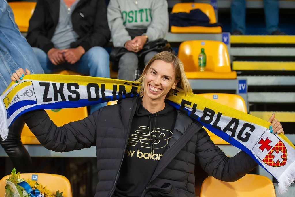Uśmiechnięta kobieta w czarnej kurtce trzyma rozwinięty szalik klubowy ZKS Olimpia Elbląg na tle żółtych trybun stadionu sportowego.