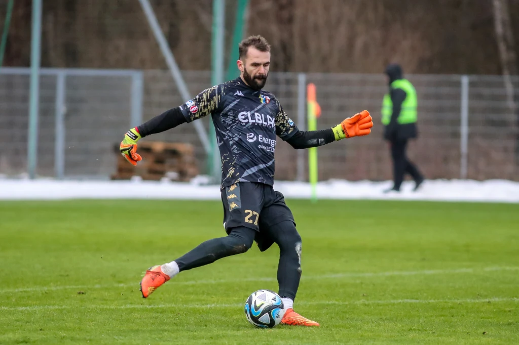 Bramkarz w ciemnym stroju kopie piłkę na zielonym boisku, w tle znajduje się sędzia liniowy oraz ogrodzenie i rozmyte drzewa.