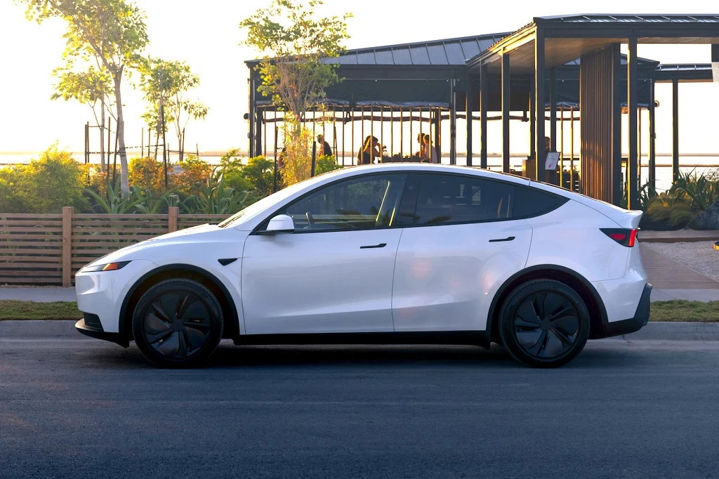 Tesla Model Y Standard Nowoczesny biały samochód elektryczny typu SUV stojący na drodze, w tle widoczne rośliny, drewniane ogrodzenie i nowoczesna altana ze szkła i metalu.
