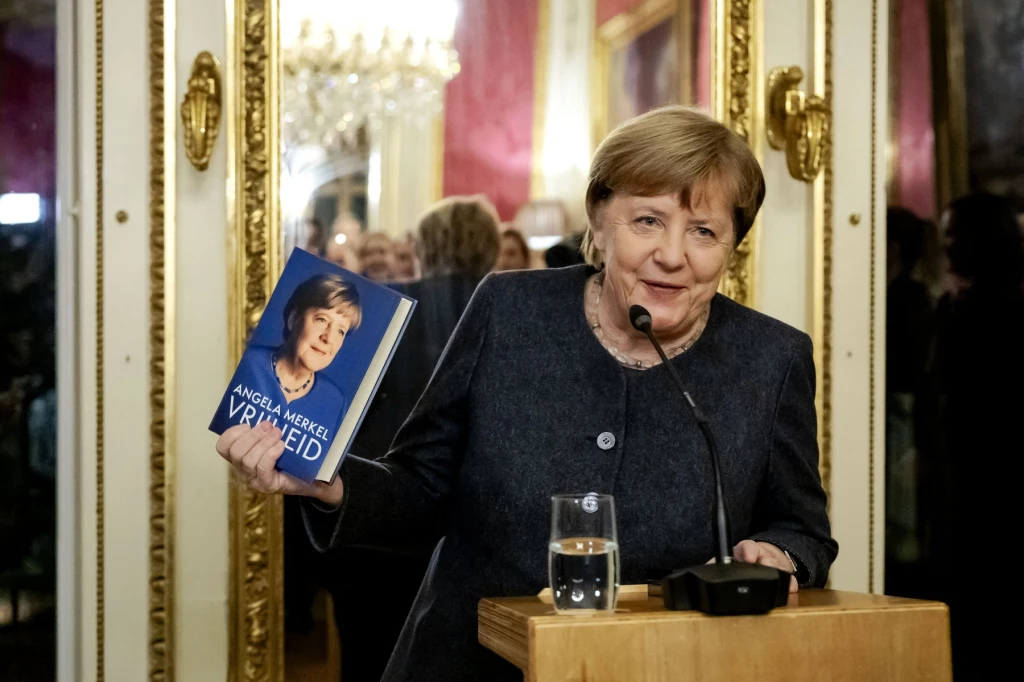 Angela Merkel ze swoją biografią "Wolność" wydaną w 2024 roku Kobieta stojąca przy mównicy, trzymająca książkę z własnym portretem na okładce, w tle pałacowe wnętrze z lustrem oraz grupą osób.