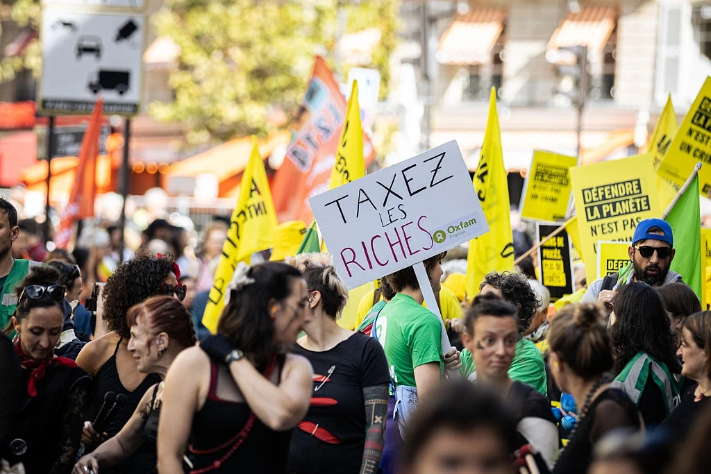 Francuzi wzywający do opodatkowania bogaczy Tłum ludzi uczestniczących w demonstracji, trzymających transparenty z hasłami dotyczącymi opodatkowania bogatych oraz obrony planety, w otoczeniu miejskiej zabudowy i roślinności.