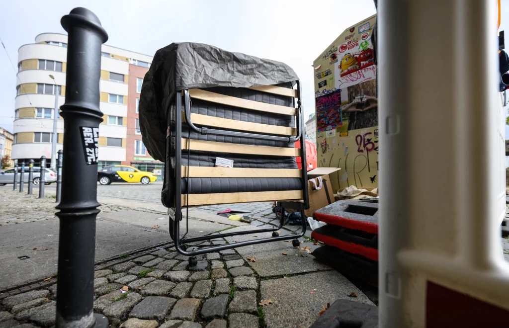 Sperrmüll to niemieckie słowo oznaczające odpady wielkogabarytowe Stary składany stelaż łóżka ustawiony na chodniku obok pojemnika oraz przedmiotów pokrytych plandeką, na tle fragmentu nowoczesnego budynku i zaparkowanego żółtego samochodu, otoczenie miejskie z elementami chodnika i betonu.