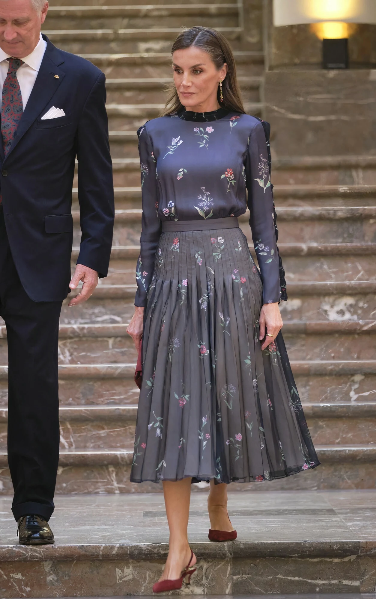 Kobieta w eleganckiej, ciemnej, wzorzystej sukience z długim rękawem i plisowaną spódnicą stoi na marmurowych schodach obok mężczyzny w garniturze, trzymając w ręce małą torebkę.