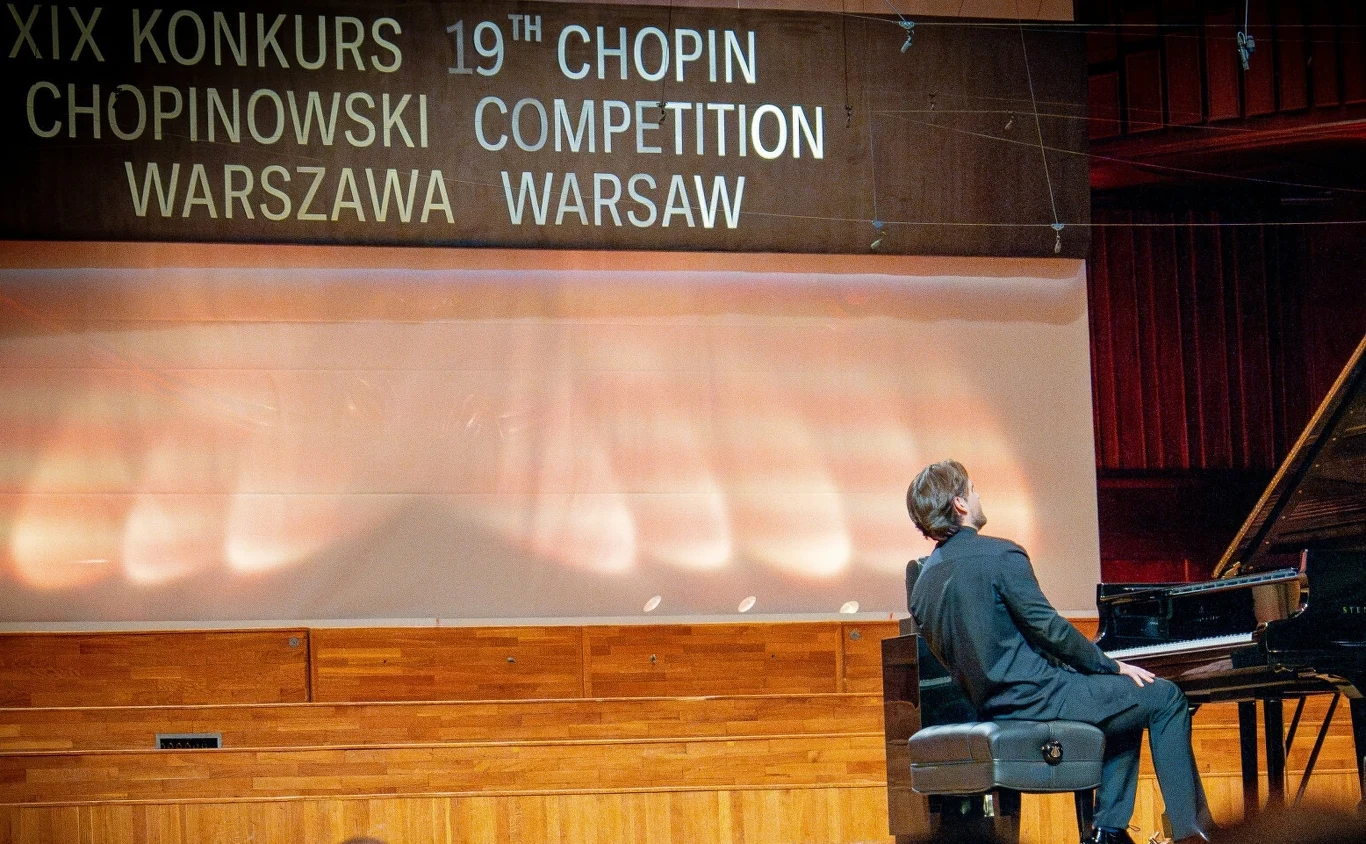 9 października odbędzie się drugi etap Konkursu Chopinowskiego Pianista siedzący przy fortepianie na scenie sali koncertowej podczas XIX Konkursu Chopinowskiego w Warszawie, nad sceną widoczne logo wydarzenia w języku polskim i angielskim, tło stanowi wnętrze sali koncertowej.