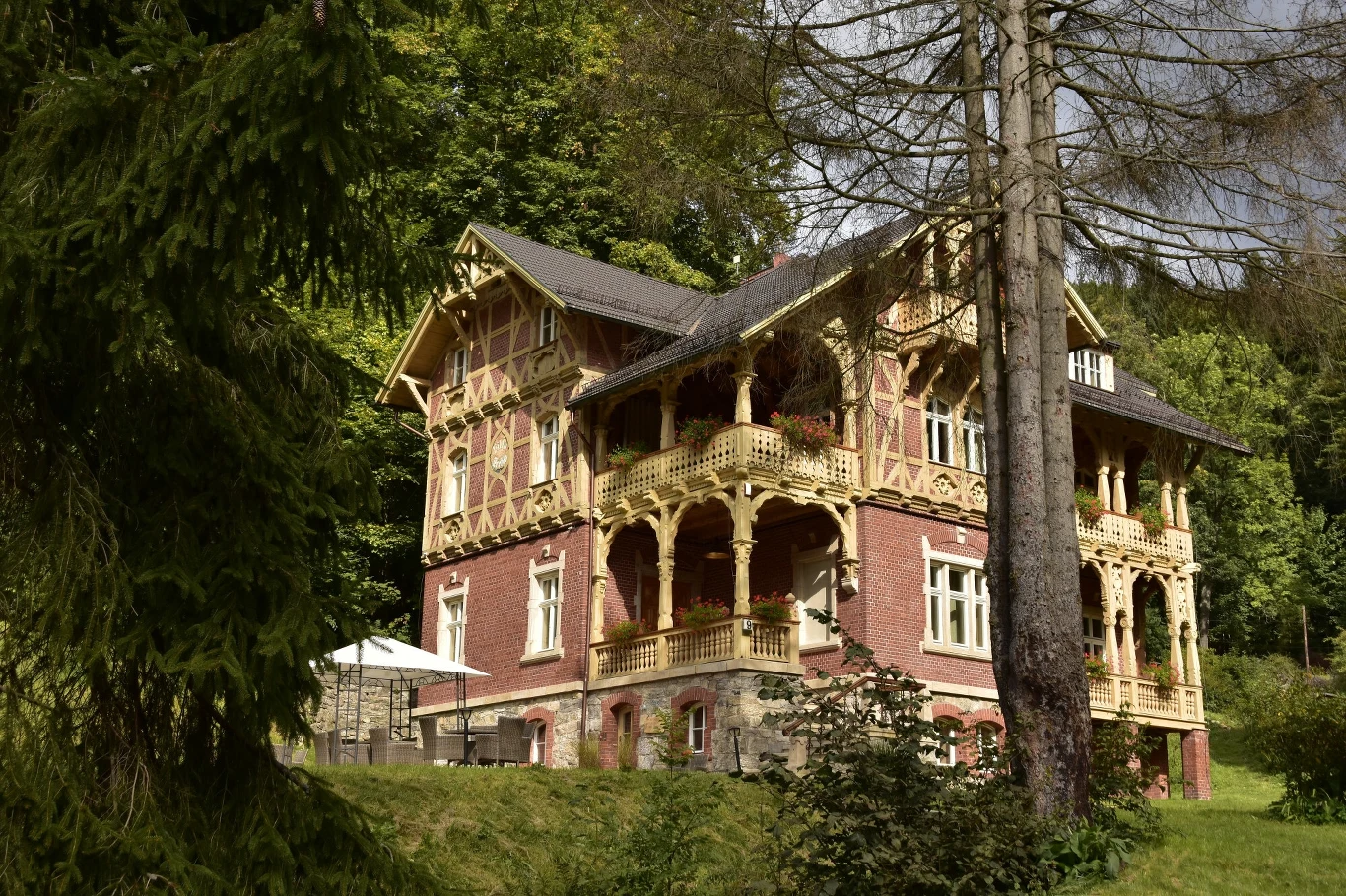 Międzygórze to architektoniczna perełka Dolnego Śląska i alternatywa dla zatłoczonych kurortów. Można tu poczuć alpejski klimat Duży, zabytkowy dom z czerwonej cegły z bogato zdobionymi drewnianymi balkonami i detalami architektonicznymi, położony wśród zielonych drzew i krzewów na lekkim wzniesieniu, na tle lasu.