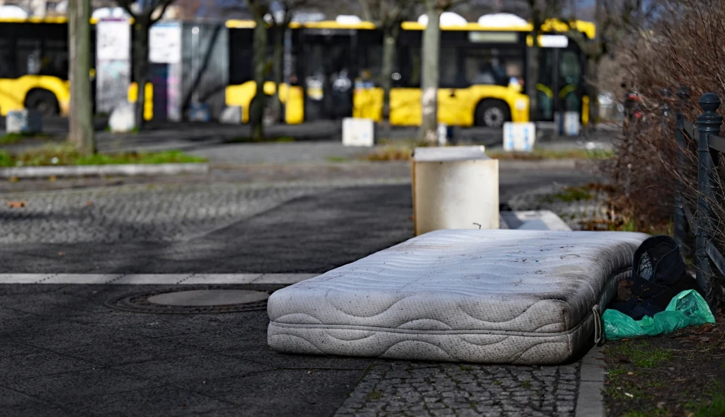 Być może wkrótce w Berlinie w życie wejdą nowe stawki kar za zostawianie przedmiotów na ulicy Porzucony materac leżący na chodniku, obok leży zielona torba i ciemny worek, w tle widoczne żółte autobusy miejskie zaparkowane przy krawędzi jezdni.
