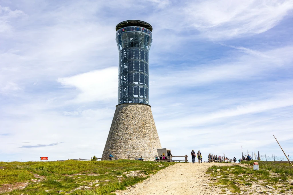 Panorama roztaczająca się ze Śnieżnika jest jeszcze bardziej imponująca dzięki nowoczesnej wieży widokowej Wieża widokowa o nowoczesnej, szklanej konstrukcji na solidnej, kamiennej podstawie, usytuowana na górskim szczycie, wokół której spacerują turyści na ścieżce oraz trawiastym terenie