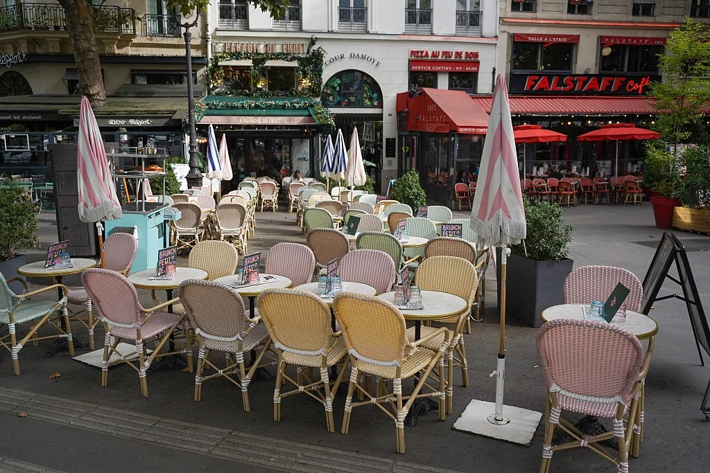 Kawiarnia w Paryżu Kolorowe krzesła i stoliki ustawione na zewnątrz kawiarni, pastelowe parasole oraz widoczne szyldy i witryny okolicznych restauracji, atmosfera typowa dla europejskich ulicznych ogródków gastronomicznych.