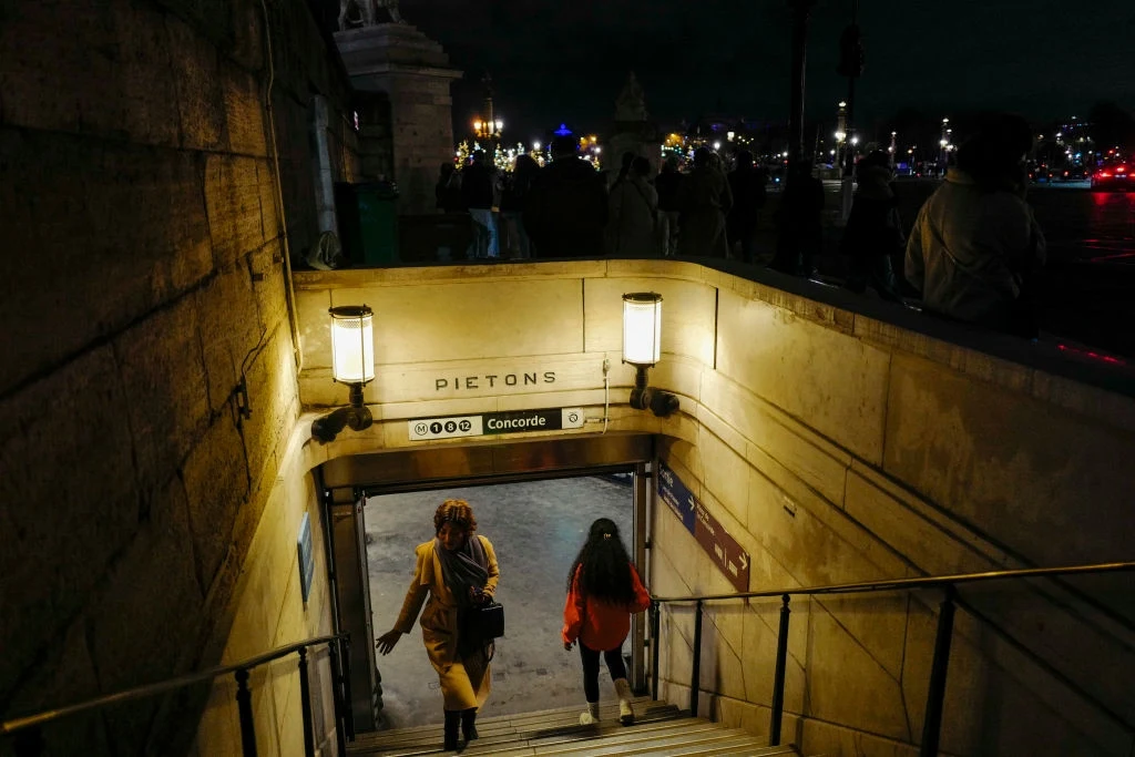 Metro w Paryżu Dwie kobiety schodzą po schodach do przejścia podziemnego oznaczonego napisem Piétons Concorde, otoczonego żółtym światłem lamp, z nocną scenerią miasta w tle.