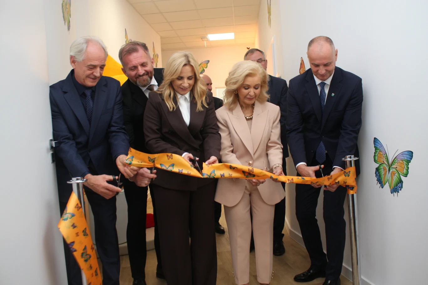 Otwarcie świetlicy w Lewinie Brzeskim Grupa elegancko ubranych osób przecina żółtą wstęgę ozdobioną motylami na korytarzu z białymi ścianami i kolorowymi motylami namalowanymi na ścianach.