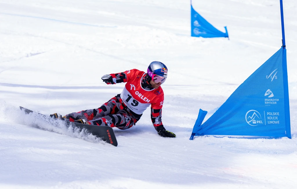 Benjamin Karl Zawodnik ubrany w czerwony strój i kask z numerem startowym 19 dynamicznie ślizga się na snowboardzie, pokonując zakręt przy niebieskiej tyczce na ośnieżonym stoku.