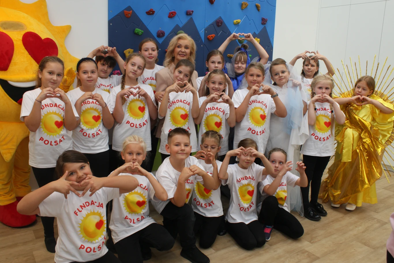 Podopieczni świetlicy w Lewinie Brzeskim Grupa dzieci i kobieta ustawieni w kilku rzędach, ubrane w białe koszulki z napisem Fundacja Polska oraz żółte elementy odzieży, wykonują gest serca dłońmi, w tle maskotka słońca i dekoracje na ścianie.