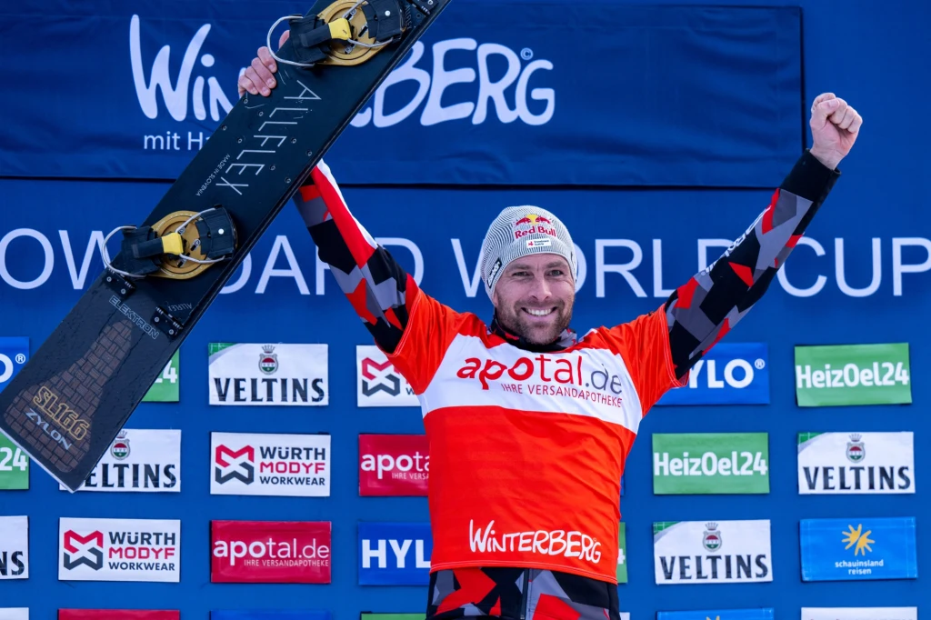 Benjamin Karl Uśmiechnięty sportowiec w stroju z numerem startowym i czapką, trzymający snowboard nad głową w geście triumfu na tle ścianki sponsorskiej, sugerującej zawody pucharu świata w snowboardzie w Winterbergu.