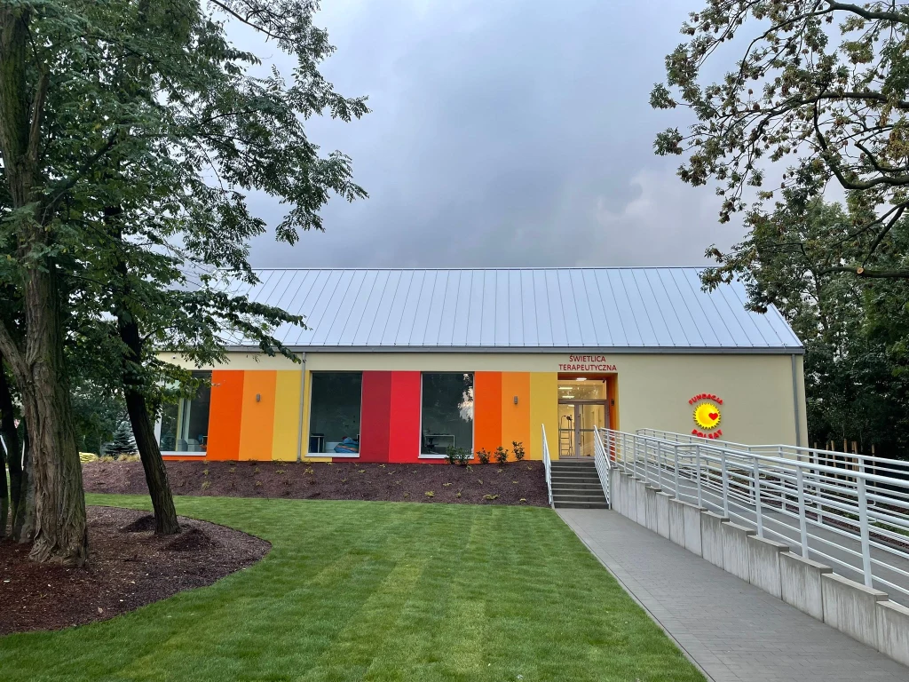 Budynek w jasnych, radosnych kolorach z elewacją ozdobioną pionowymi pasami żółci, czerwieni i pomarańczu, szeroka ścieżka prowadzi do wejścia, wokół świeżo przycięty trawnik i drzewa, po prawej stronie napis Klub Terapeutyczny i symbol słoneczka.