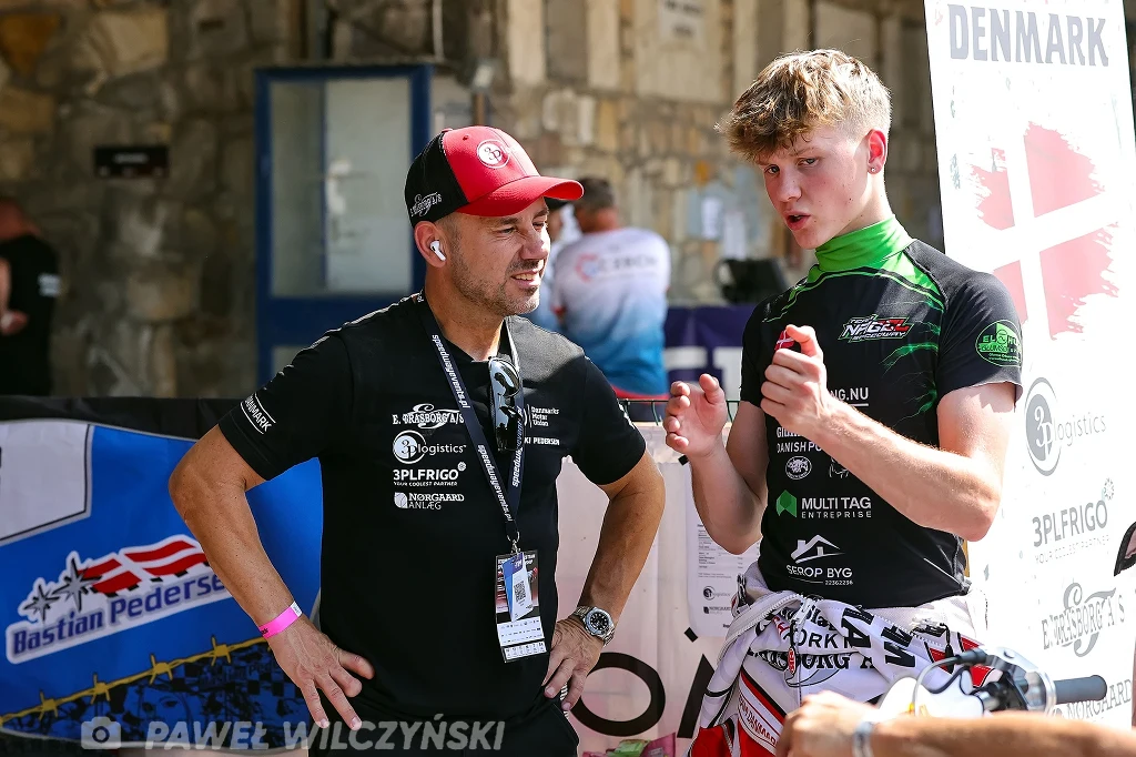 Dwóch mężczyzn rozmawia ze sobą podczas wydarzenia sportowego, jeden z nich nosi czarną koszulkę i czerwoną czapkę z daszkiem, drugi ubrany jest w sportowy strój z zielonym akcentem, w tle widoczne są banery z logo oraz napisem Denmark.