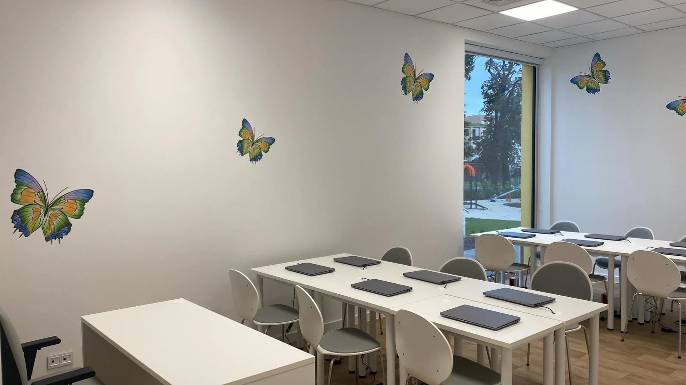 Wnętrze Świetlicy Terapeutycznej w Lewinie Brzeskim Jasna sala z białymi ścianami ozdobionymi kolorowymi motylami, kilka białych stołów z ustawionymi tabletami oraz szare krzesła, duże okno z widokiem na zieleń na zewnątrz.