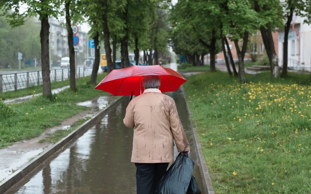 Starsza osoba idąca chodnikiem wzdłuż mokrej alejki, trzymająca czerwoną parasolkę oraz czarną torbę na tle drzew i wiosennej zieleni po deszczu.