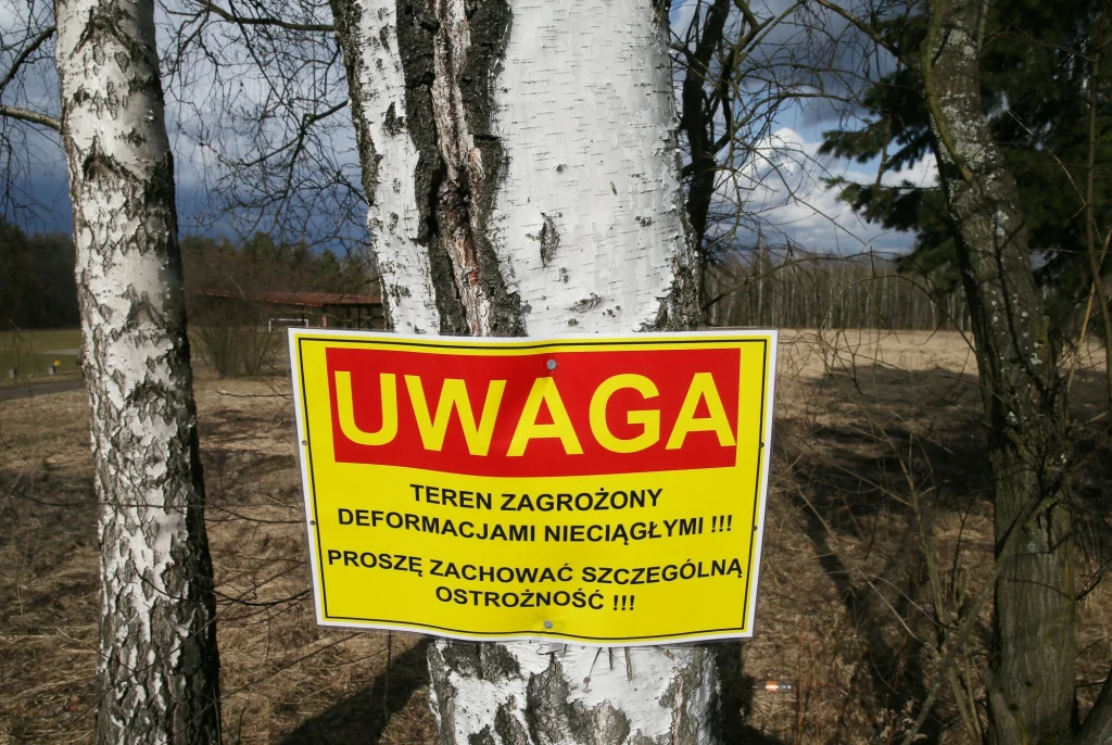 Jasna tablica ostrzegawcza z czerwonym napisem UWAGA umieszczona na pniu brzozy, informująca o zagrożeniu deformacjami terenu i apelująca o zachowanie szczególnej ostrożności, w tle widać drzewa i nieużytki.
