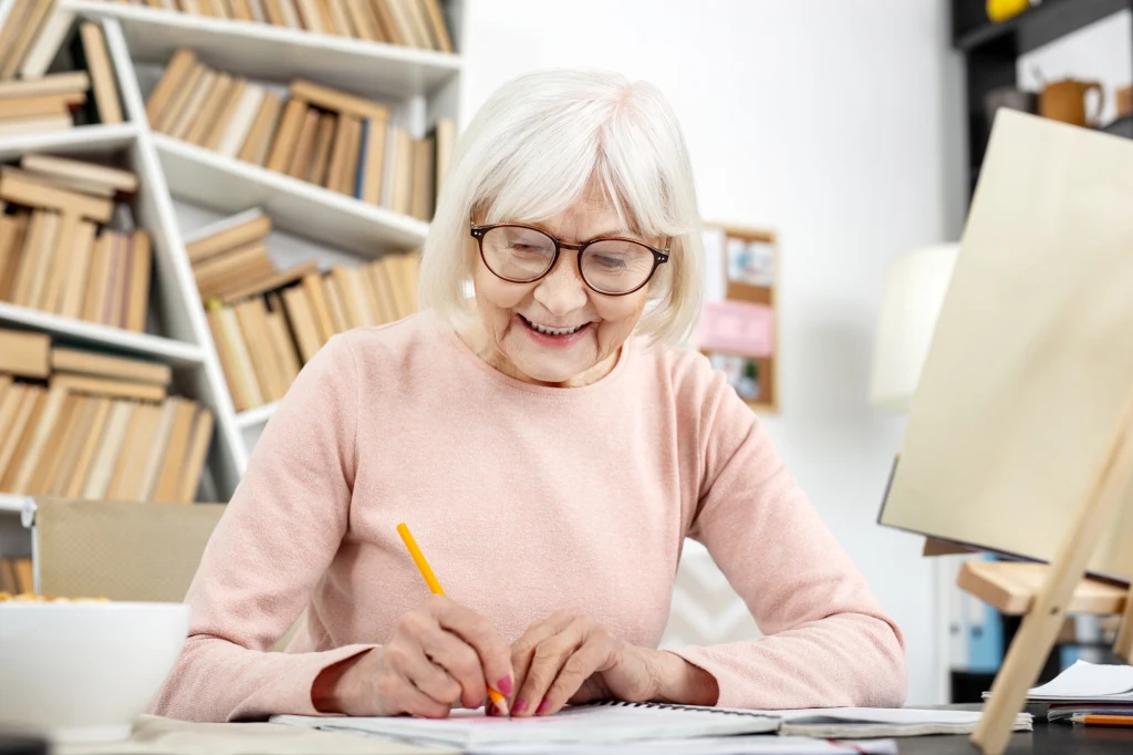 Nie przestawaj się uczyć, bez względu na wiek Starsza kobieta z siwymi włosami i okularami pisze przy biurku, w tle regał z licznymi książkami oraz komputer, tworząc atmosferę skupienia i nauki.