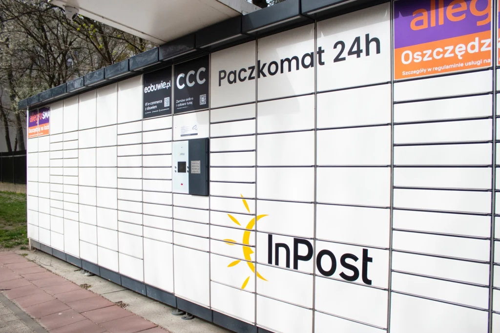 Możesz postawić paczkomat na swojej działce. Warunki są proste Automatyczny paczkomat firmy InPost z licznymi skrytkami na paczki oraz panelami do obsługi przesyłek, widoczne reklamy sklepów internetowych oraz oznaczenie działania 24h.