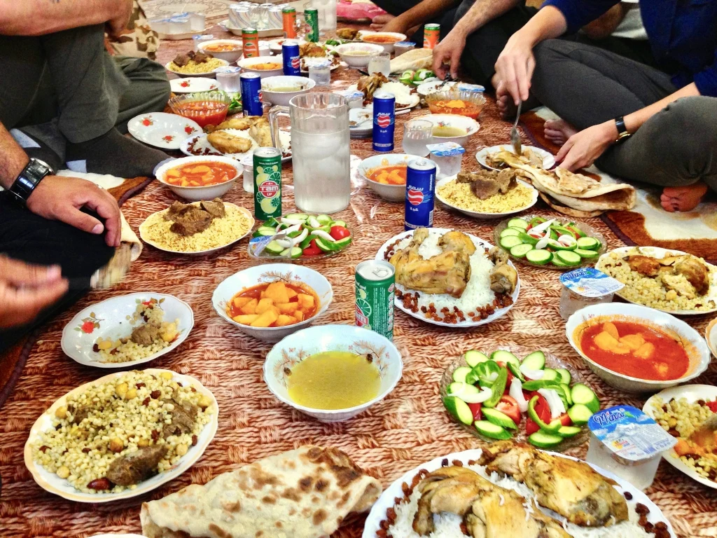 Ręce kilku osób rozkładających posiłek na tradycyjnych talerzach, dużo potraw w typie kuchni bliskowschodniej, różnorodne dania z warzyw, mięsa, ryżu, pieczywo, talerze sałatki, napoje w puszkach na materiale pełniącym rolę obrusa.