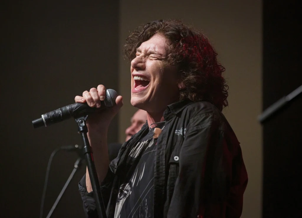 Eric Martin (Mr. Big) - Berlin, 12 kwietnia 2024 r. Piosenkarz z długimi kręconymi włosami śpiewa do mikrofonu z intensywną ekspresją twarzy, na scenie o ciemnym tle, w tle widoczna sylwetka drugiej osoby.