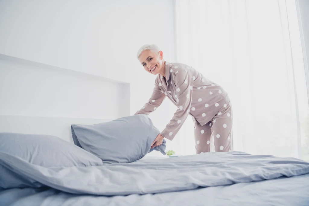 Ogranicz ekrany przed położeniem się spać Starsza kobieta z krótkimi siwymi włosami w piżamie w grochy ścieli łóżko w jasnej, schludnej sypialni, uśmiechając się podczas układania poduszek.