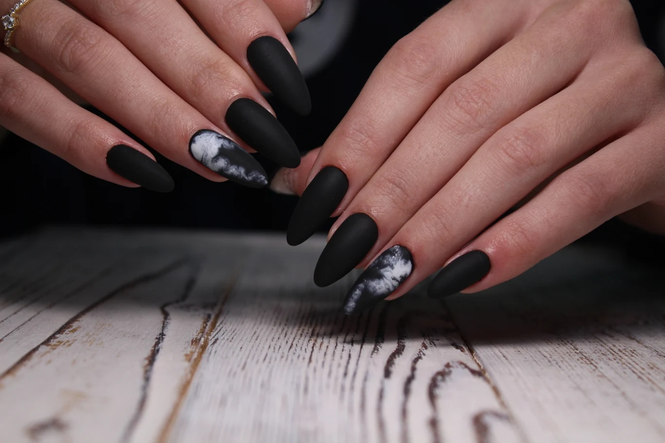 Mroczne motywy na paznokcie na Halloween 2025? Zobacz co jest teraz w trendach Dłonie z długimi, czarnymi matowymi paznokciami, część paznokci ozdobiona białymi malunkami przypominającymi marmur na tle jasnego, drewnianego stołu.
