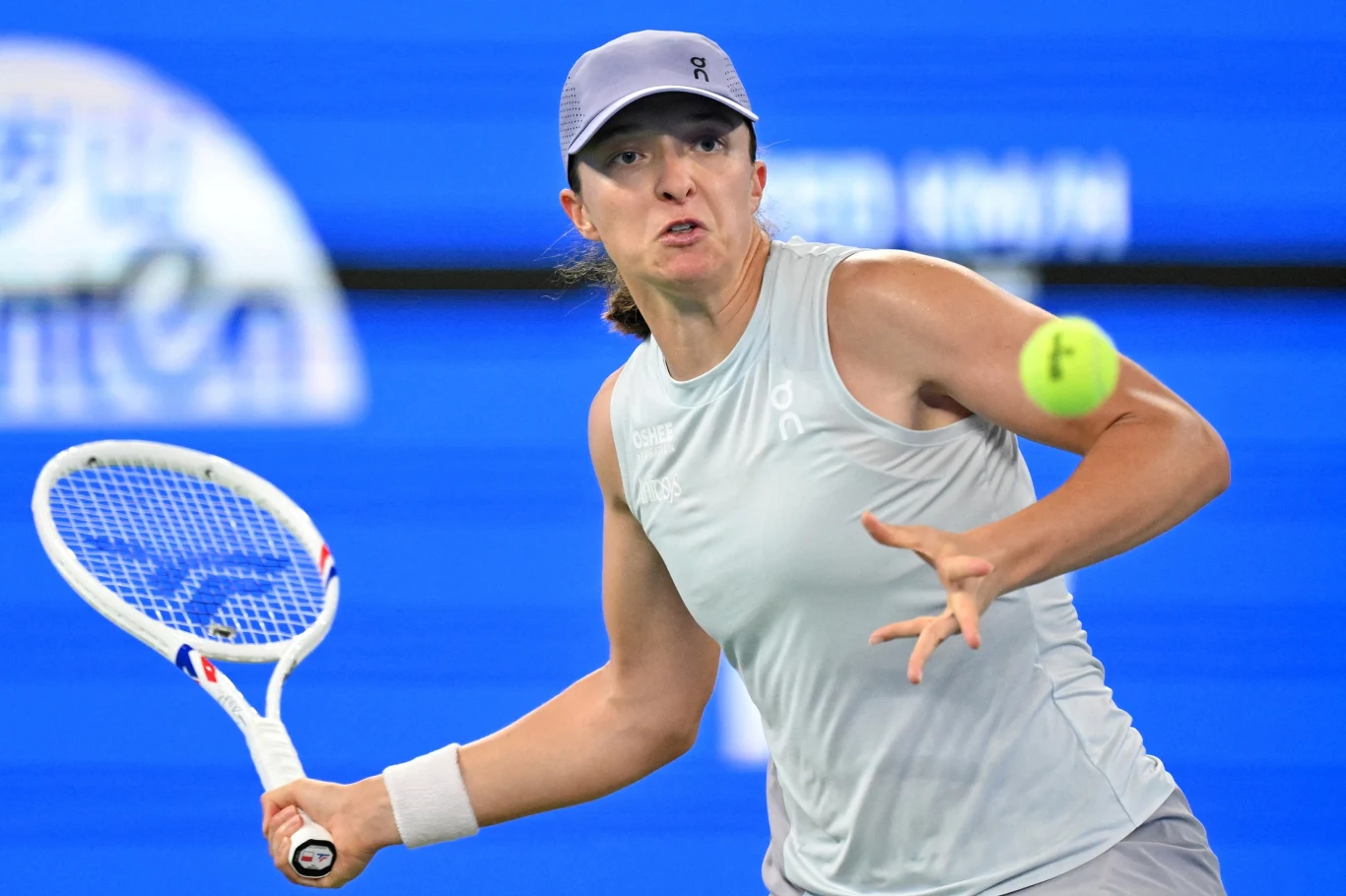 Iga Świątek podczas turnieju WTA 1000 w Wuhan 2025 Zawodowa tenisistka w jasnej sportowej koszulce i czapce z daszkiem wykonuje dynamiczny ruch, uderzając piłkę tenisową rakietą podczas meczu na tle niebieskiego kortu.
