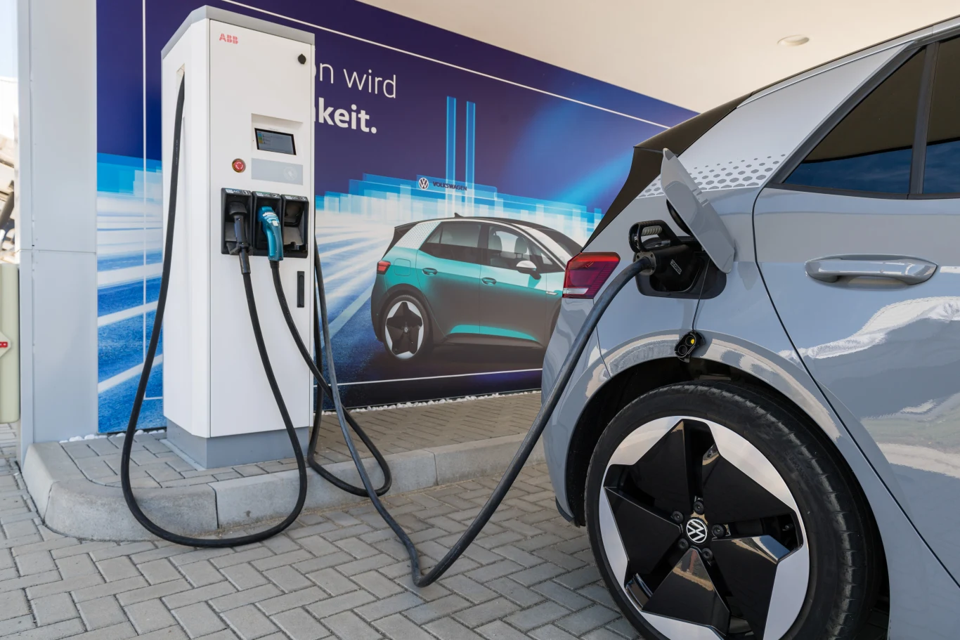 Czy samochody elektryczne powstają z etycznie pozyskiwanych surowców? Nowoczesny szary samochód elektryczny marki Volkswagen podłączony do białej stacji ładowania na tle plakatu promującego elektromobilność.