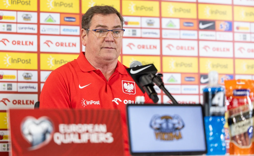 Jan Urban Trener reprezentacji Polski w piłce nożnej siedzi przy stole podczas konferencji prasowej, w tle plansza z logotypami sponsorów, na stole mikrofon oraz napoje sportowe.