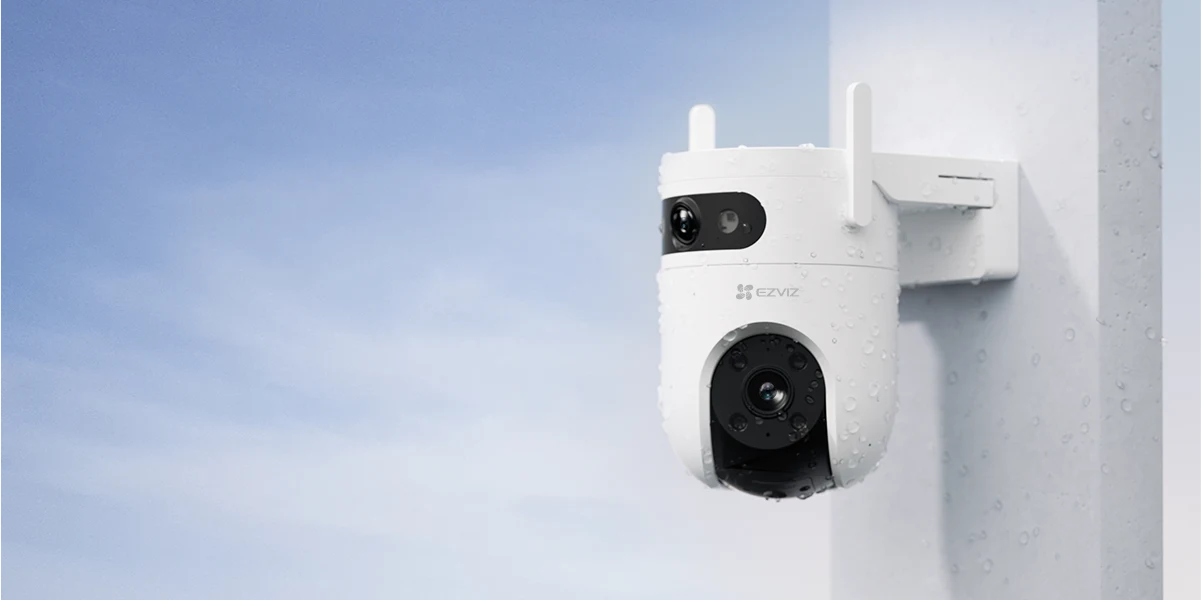 EZVIZ H9C wygląda elegancko Biała kamera monitoringu zewnętrznego zamontowana na jasnej ścianie budynku, delikatnie pokryta kroplami wody, na tle niebieskiego, lekko zachmurzonego nieba.