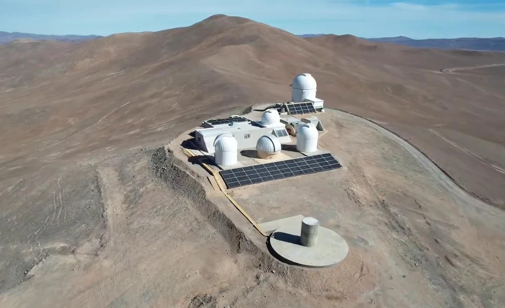 Kompleks obserwatorium astronomicznego z charakterystycznymi kopułami na szczycie wzgórza, otoczony panelami słonecznymi i położony w suchym, górzystym terenie.