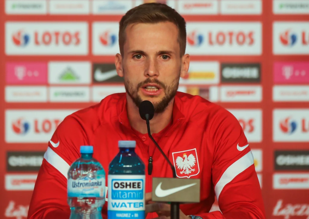 Piłkarz w czerwonym dresie reprezentacji Polski siedzi przy stole konferencyjnym z mikrofonem, za nim ścianka sponsorów, na stole butelki z wodą i inne przedmioty.