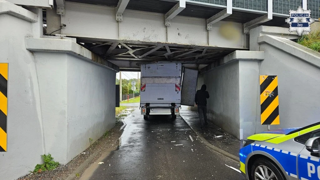 Zabudowa samochodu została uszkodzona Dostawczy samochód zaklinowany pod niskim wiaduktem kolejowym, rozbity tył pojazdu, stojąca obok osoba oraz radiowóz policyjny z przodu po prawej stronie.