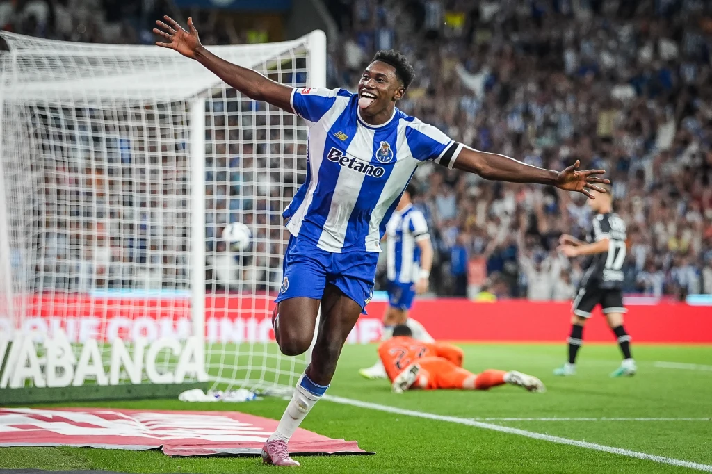 Samu Aghehowa Piłkarz drużyny FC Porto z szeroko rozłożonymi rękami cieszy się po zdobytej bramce, w tle bramkarz i inny zawodnik drużyny przeciwnej znajdują się na murawie, liczna publiczność dopingująca z trybun.