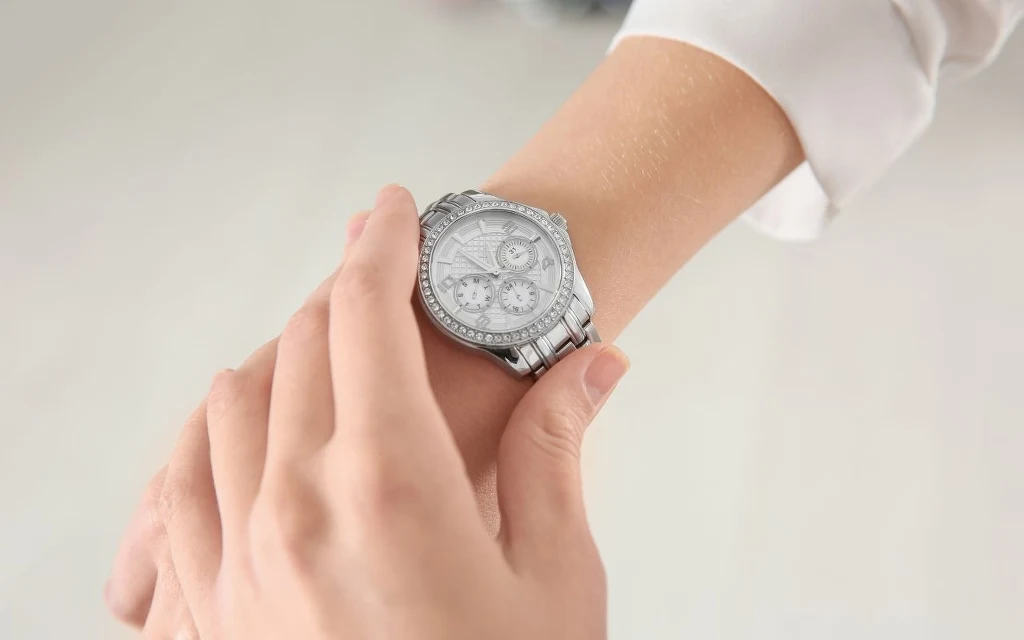 Wybierasz się na cmentarz? Zdejmij zegarek z ręki Srebrny zegarek na ręku osoby ubrane w białą koszulę, druga dłoń dotyka zegarka, tło rozmyte, jasne.